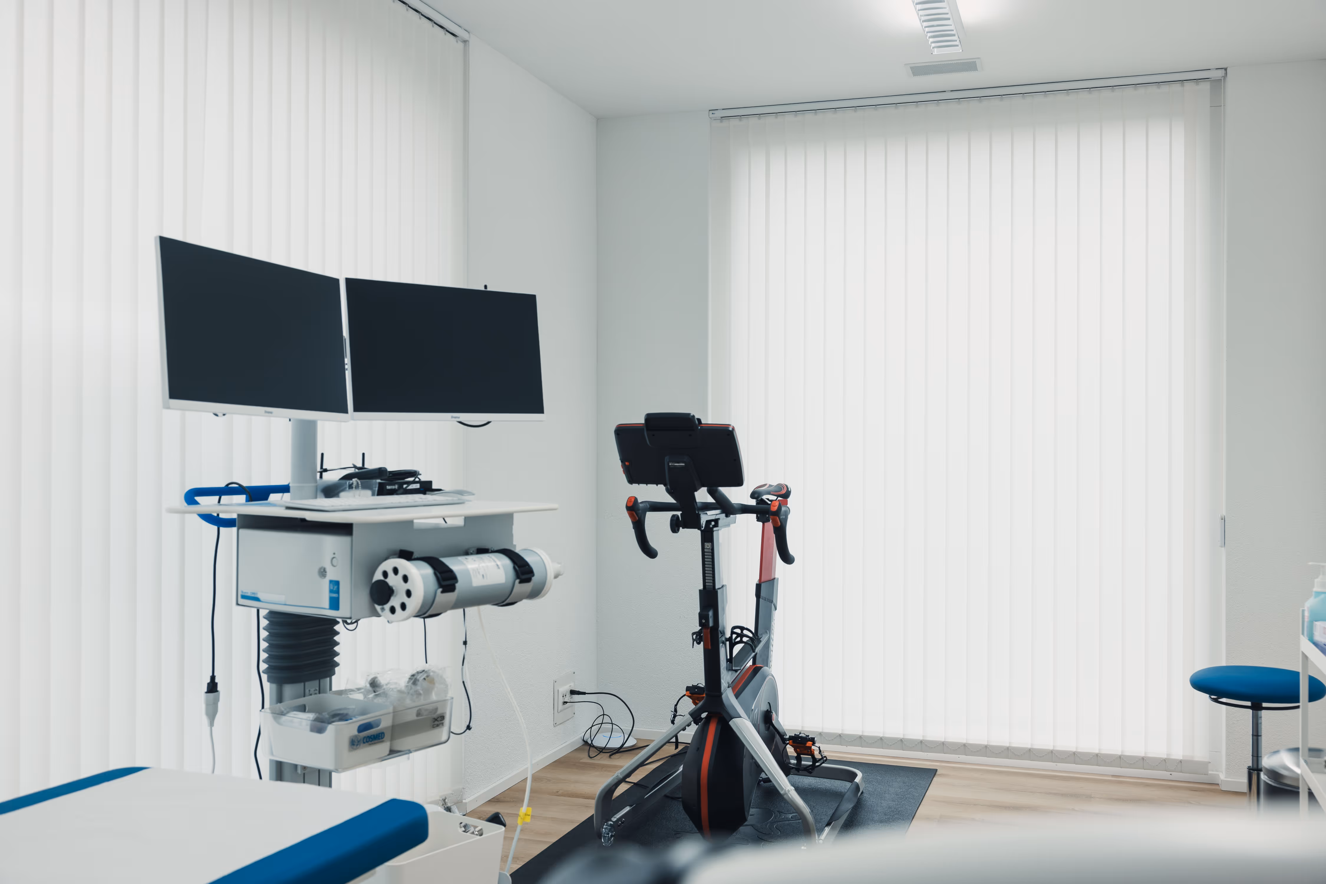 Medical examination room with a stationary exercise bike, dual computer monitors on a desk, and medical supplies near large vertical blinds-covered windows.