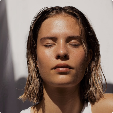 Close-up of a young woman with wet hair and closed eyes, softly lit, against a neutral background.