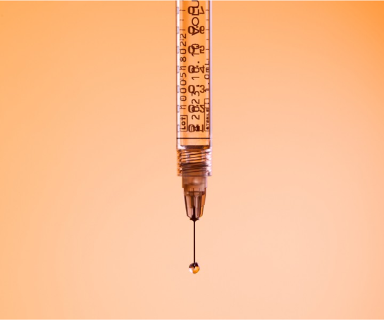 Close-up of a syringe with a droplet at the tip of the needle against an orange background.