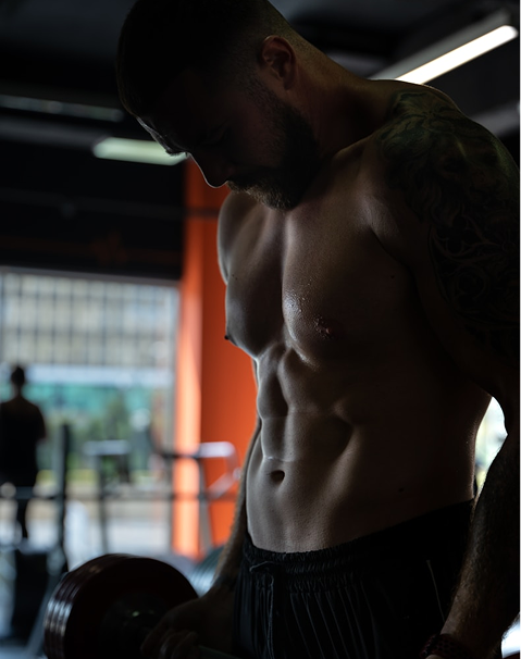 Muscular man with a tattoo lifting a dumbbell in a dimly lit gym.