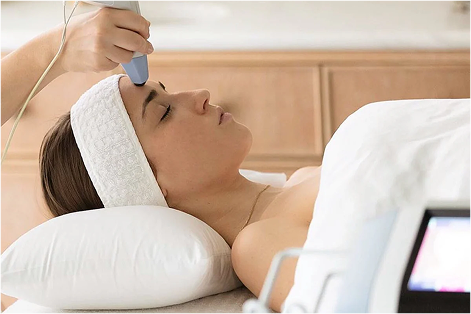 Woman lying on a pillow with eyes closed receiving a facial treatment using a handheld device on her forehead.
