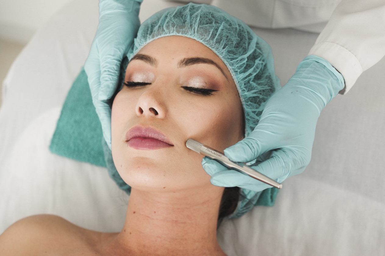 A woman with closed eyes receiving a facial treatment using a metal tool by a gloved professional wearing a hair net.