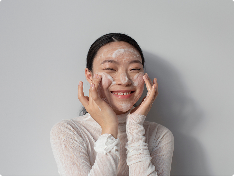 Smiling woman applying foaming facial cleanser to her face against a light gray background.