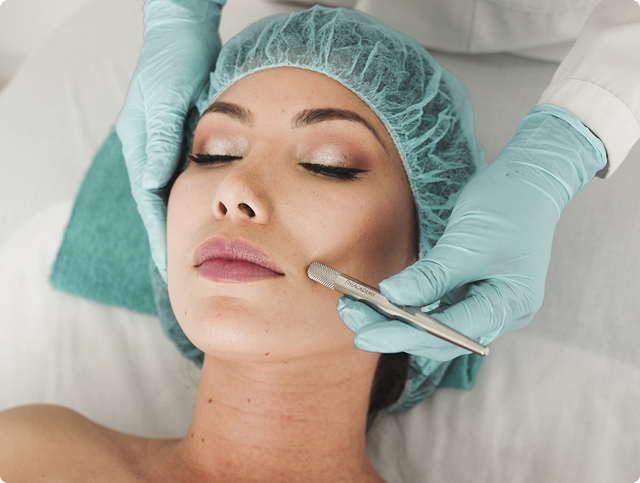 Woman receiving a facial treatment with a handheld metal tool, wearing a blue hair cap and lying on a treatment bed.