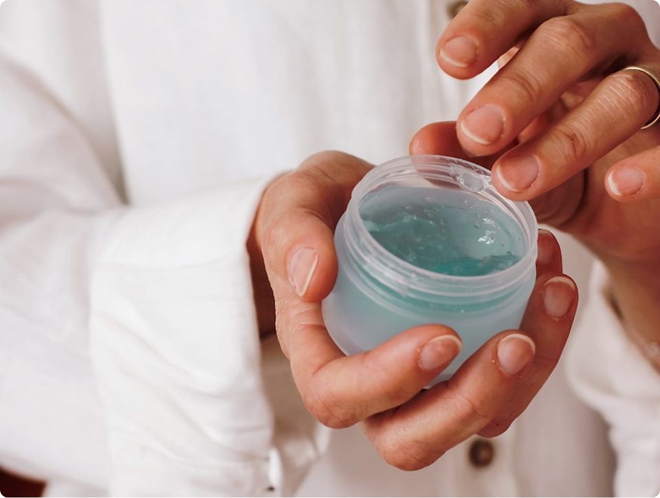 Person wearing a white shirt holding a small jar of blue gel skincare cream and dipping a finger into it.