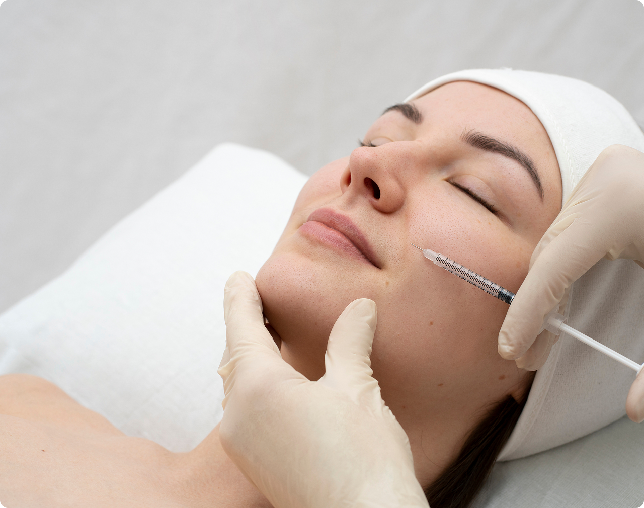 Close-up of a woman receiving a cosmetic injection in her cheek while lying down with eyes closed.