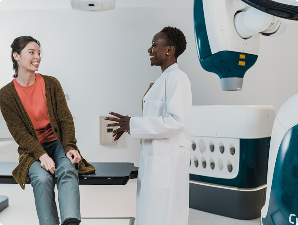 Doctor in a white coat smiling and talking with a female patient sitting on a medical examination table in a clinical room with medical equipment.