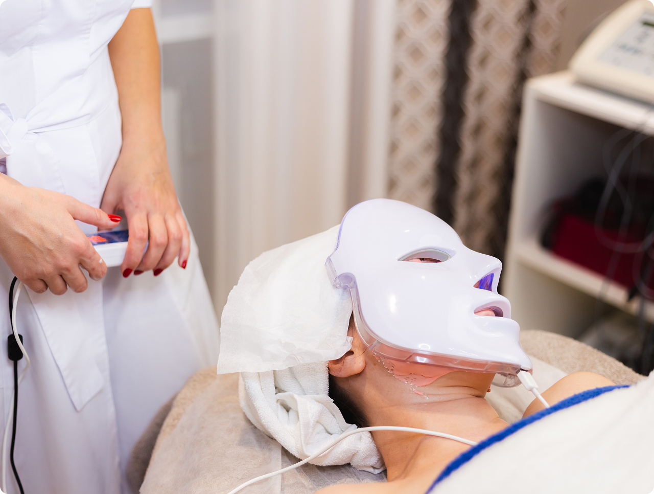 Person lying on a spa bed wearing a white LED light therapy facial mask, with a practitioner in white holding a control device.