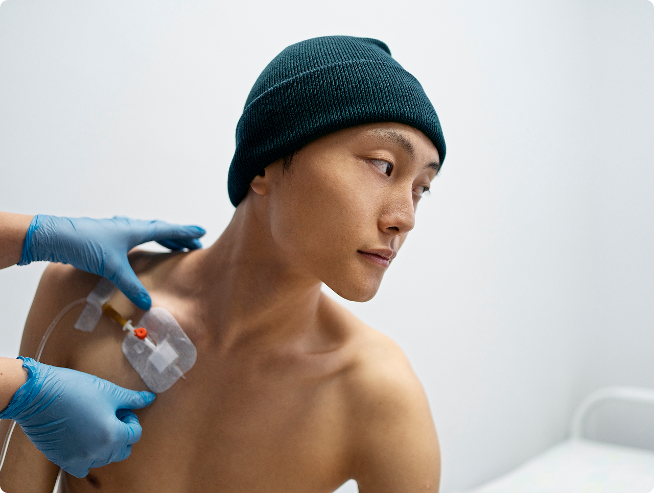 Medical professional wearing blue gloves adjusting a catheter on a shirtless man wearing a dark beanie.