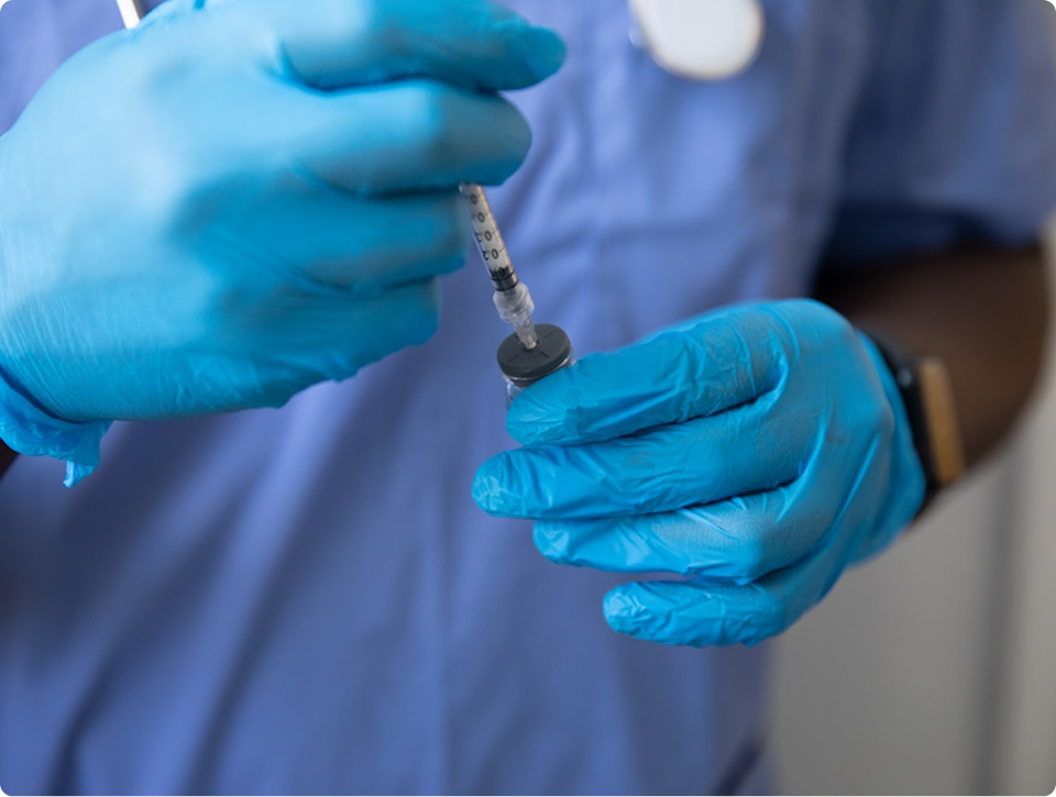 Healthcare worker wearing blue gloves filling a syringe with a vaccine from a vial.