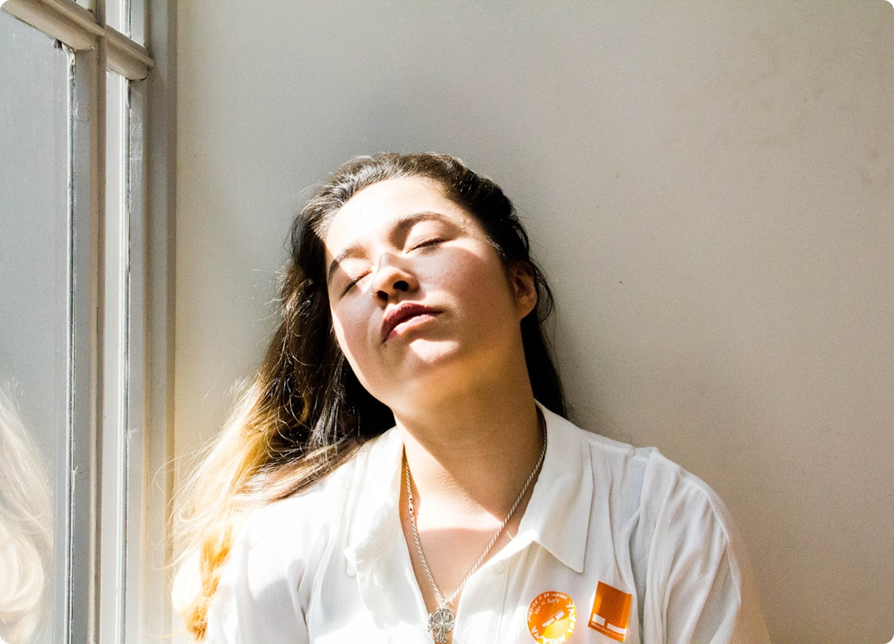 Woman leaning against a wall near a window with sunlight on her face and eyes closed.