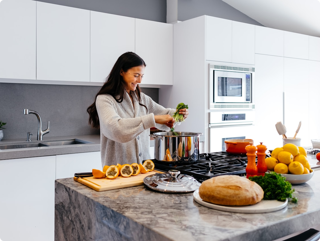 Woman adding greens to a cooking pot in a modern kitchen with fresh ingredients on the counter including papaya halves, lemons, bread, and herbs.