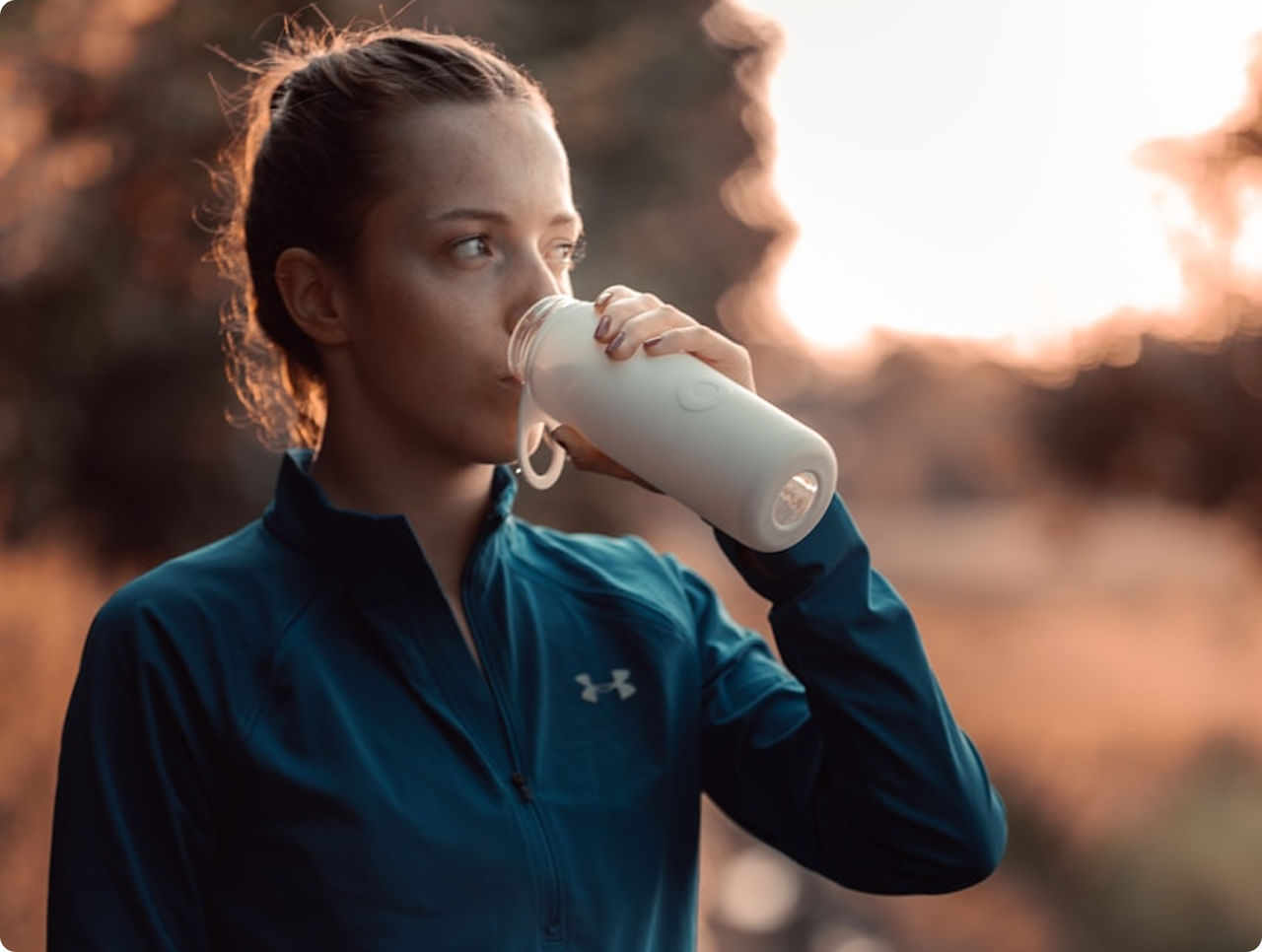Woman in blue athletic jacket drinking from a white water bottle outdoors at sunset.