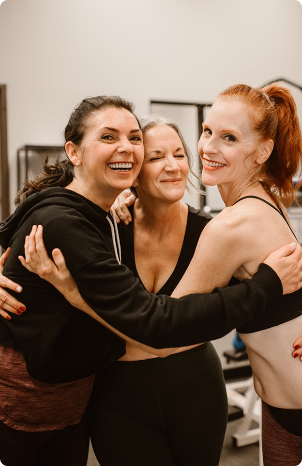 Three women in workout clothes smiling and hugging each other in a gym setting.