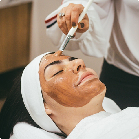 Woman with eyes closed receiving a brown facial mask applied with a brush by an esthetician.