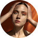 Close-up of a woman with natural makeup holding her face with both hands against an orange background.