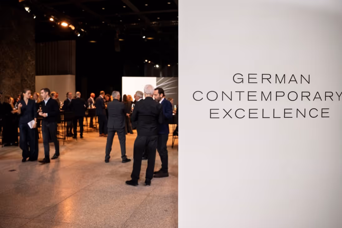 People socializing in a modern event space next to a wall displaying 'German Contemporary Excellence' text.