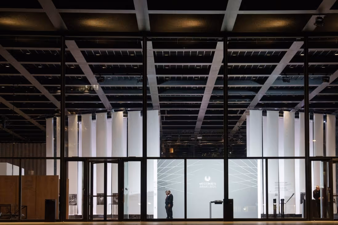 Night view through large glass panels showing two people standing inside a modern, brightly lit building with vertical white columns.