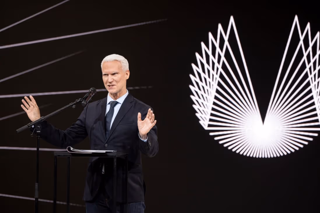 Man in suit speaking at a microphone with geometric light pattern on black background.