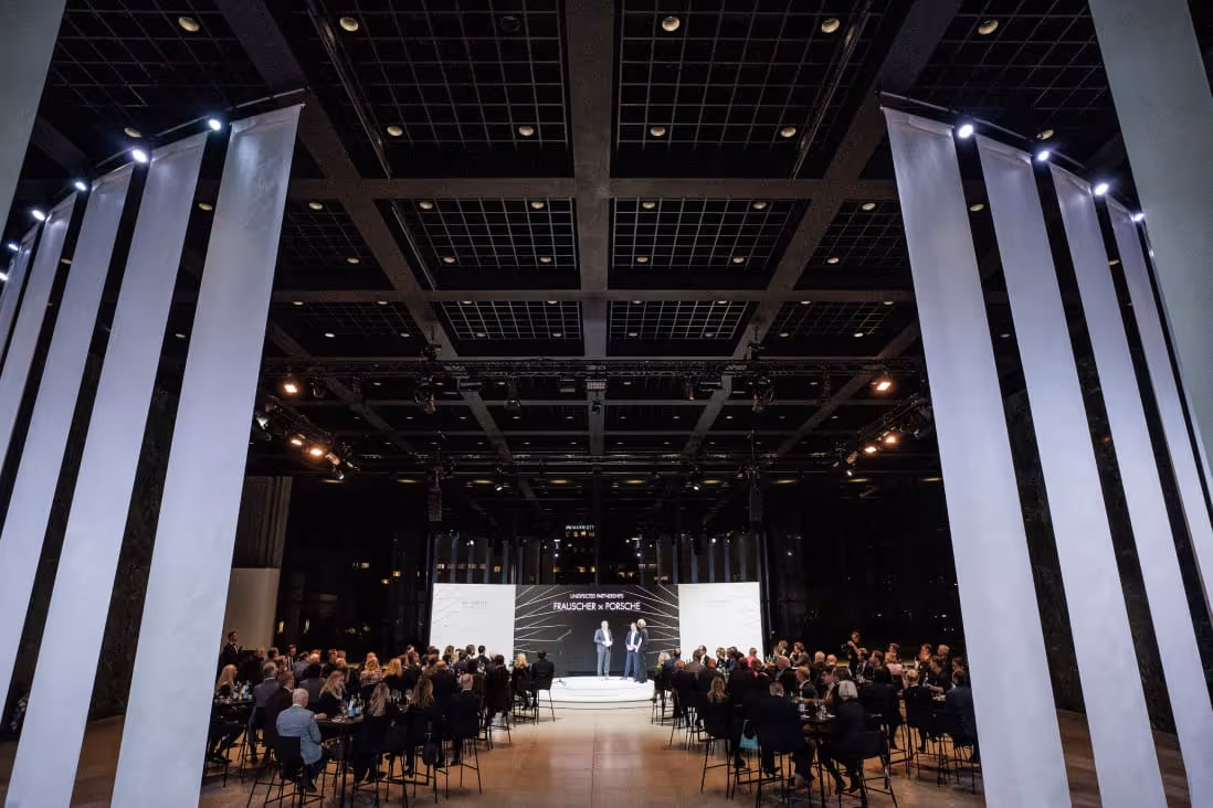 Large indoor event with many seated attendees facing a stage where two people are speaking under a banner that reads 'UNEXPECTED MEETS FRAUSCHER x PORSCHE.'