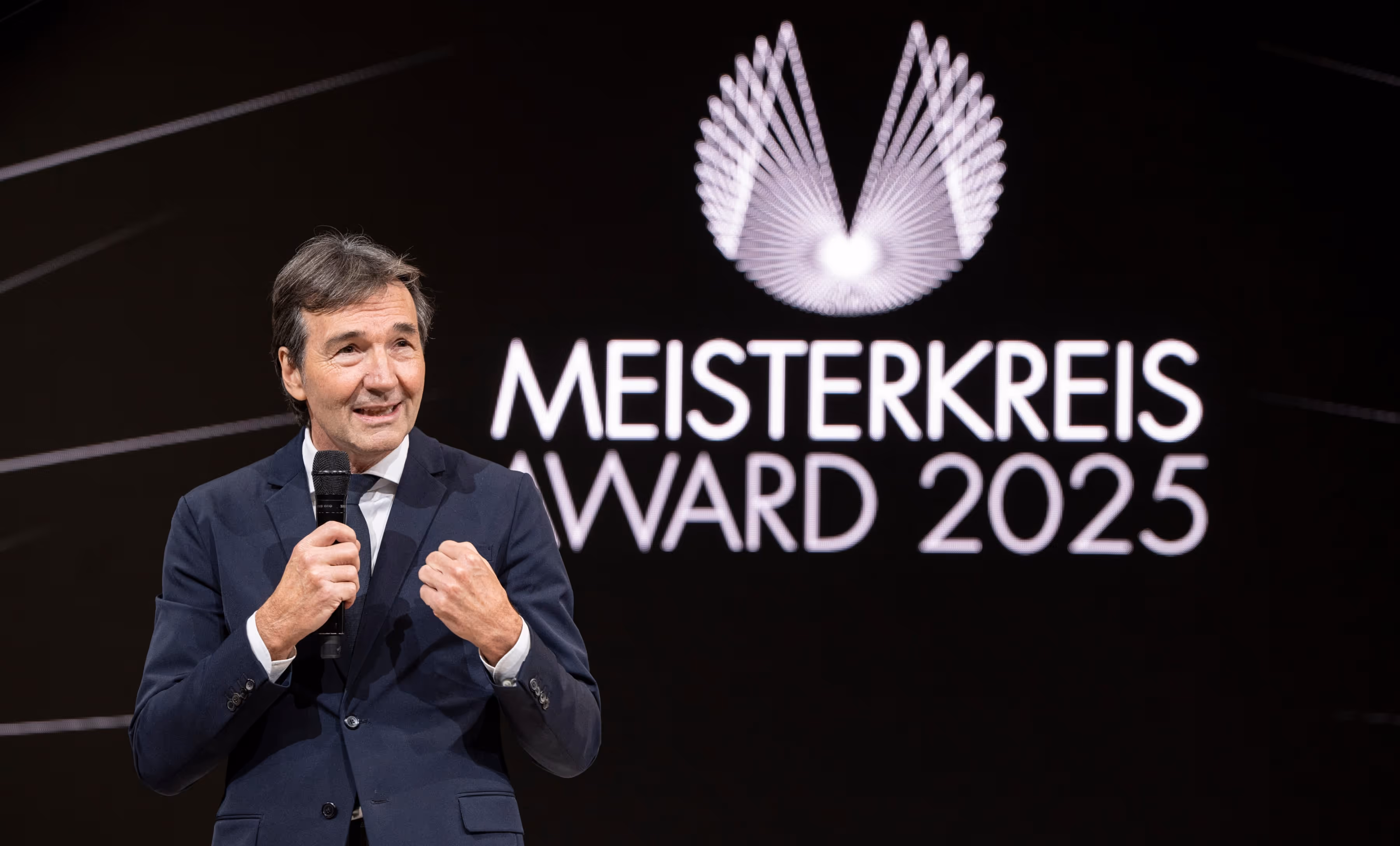 Man in a dark suit holding a microphone speaking at the Meisterkreis Award 2025 event with event logo in the background.