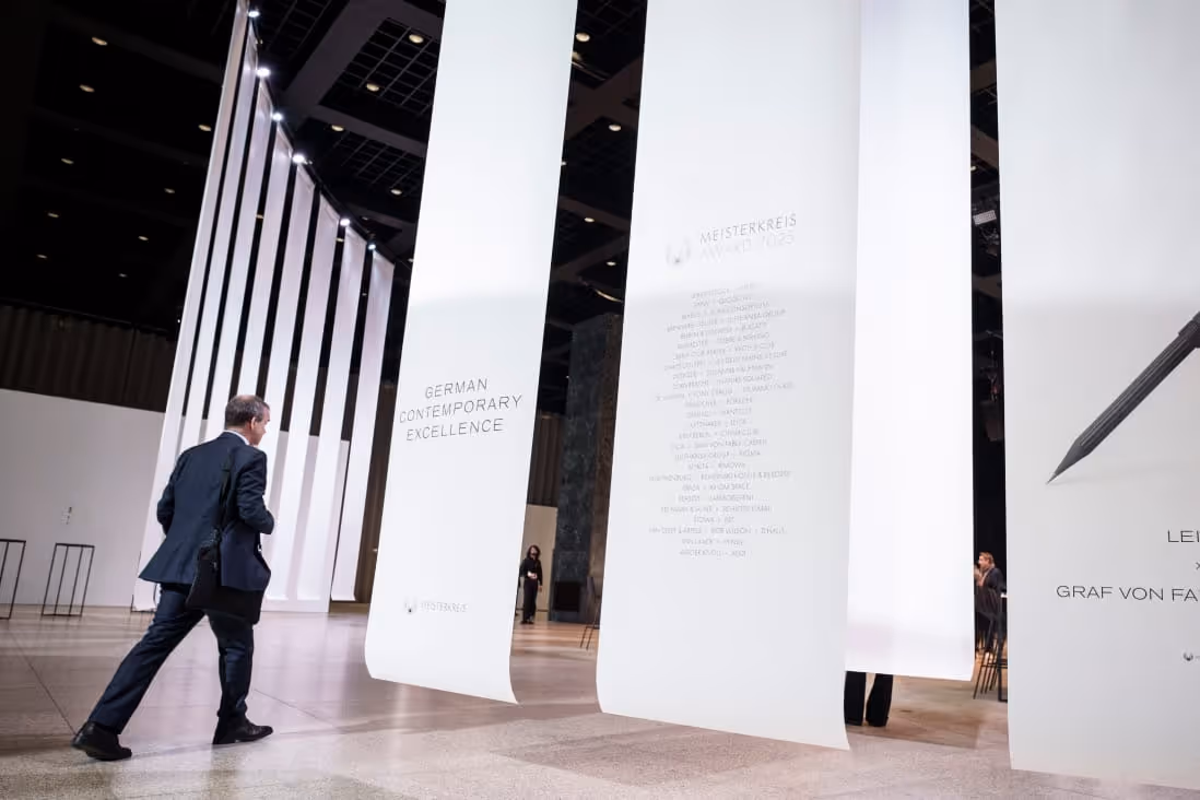 A man in a suit walks through an exhibition with large white vertical banners displaying text about German contemporary excellence and the Meisterkreis Award 2025.