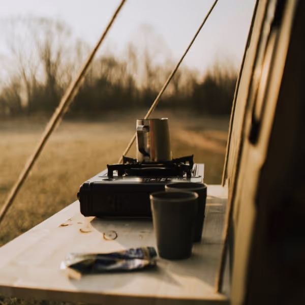 Campingkocher mit Metallbecher auf einem Campingtisch neben zwei Bechern und einer Zeltwand im Hintergrund eines offenen Feldes.