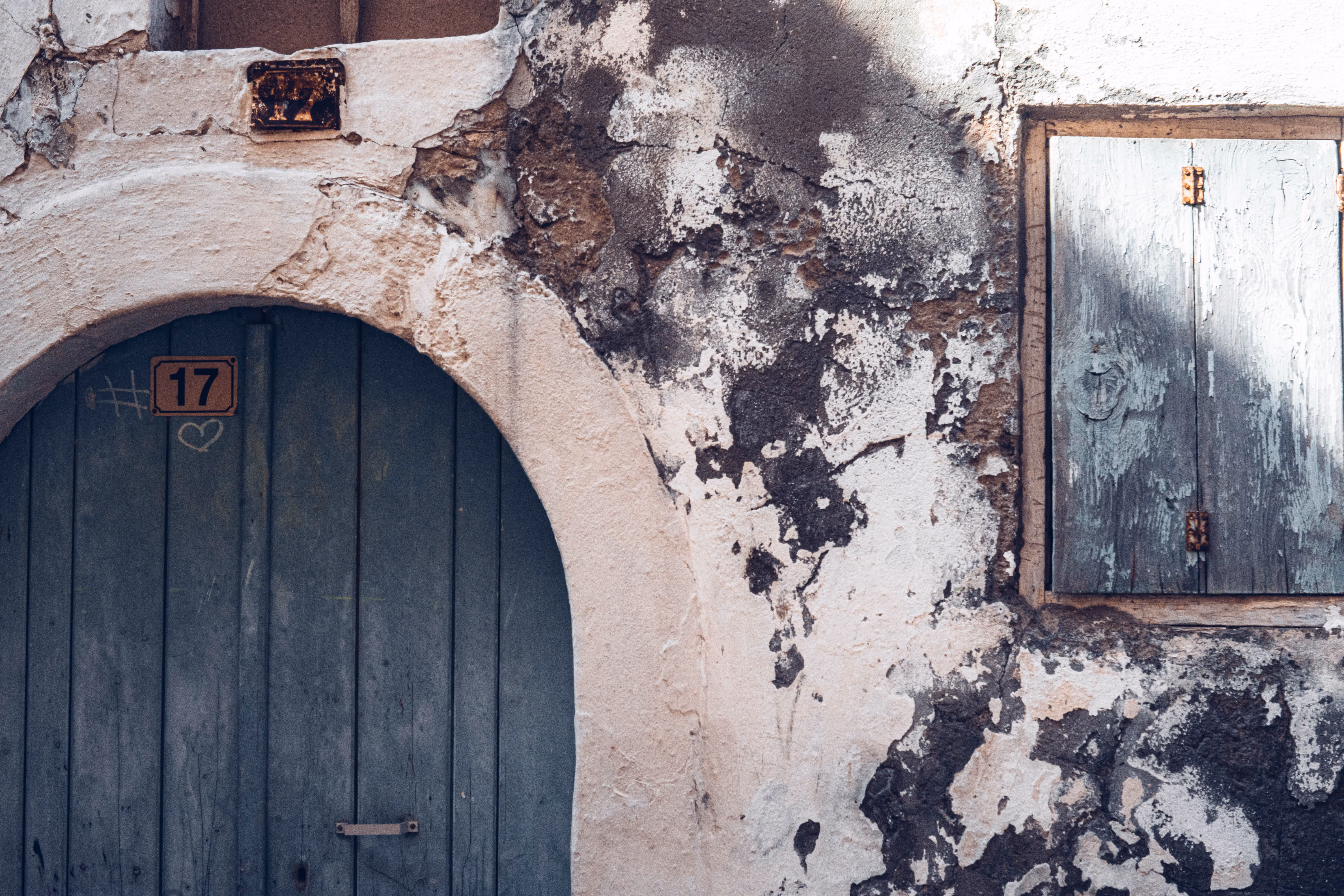 Verwitterte Wand mit blauem Holztor und Fenster mit verschlossenen grünen Holzläden.