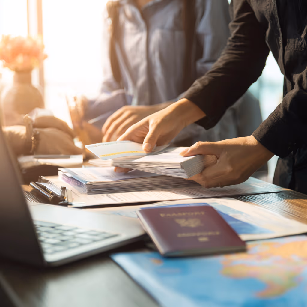Handen die een stapel vliegtickets vasthouden op een bureau met een laptop, paspoort en wereldkaart.