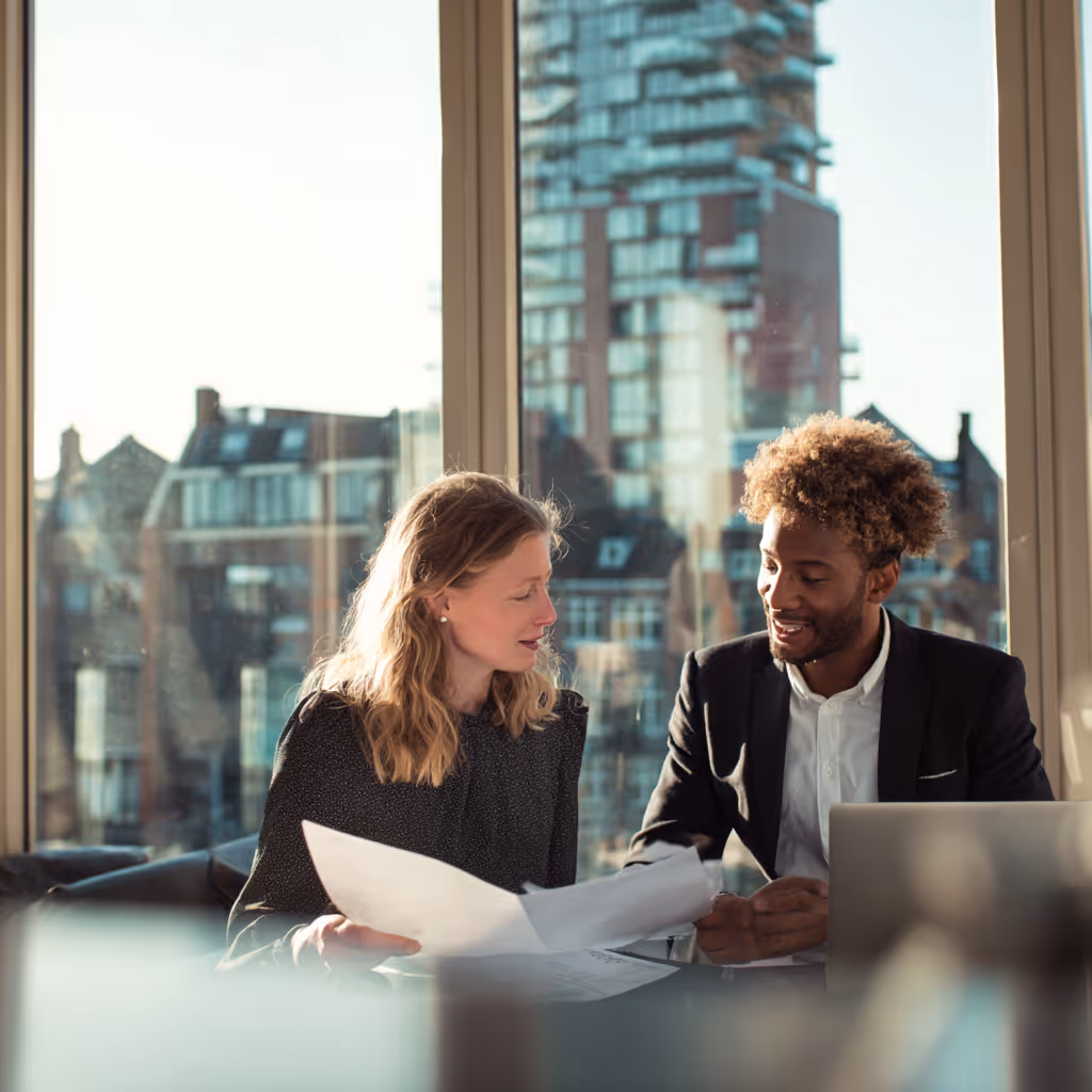 Twee professionals zitten aan tafel in een kantoor met grote ramen en bespreken documenten.