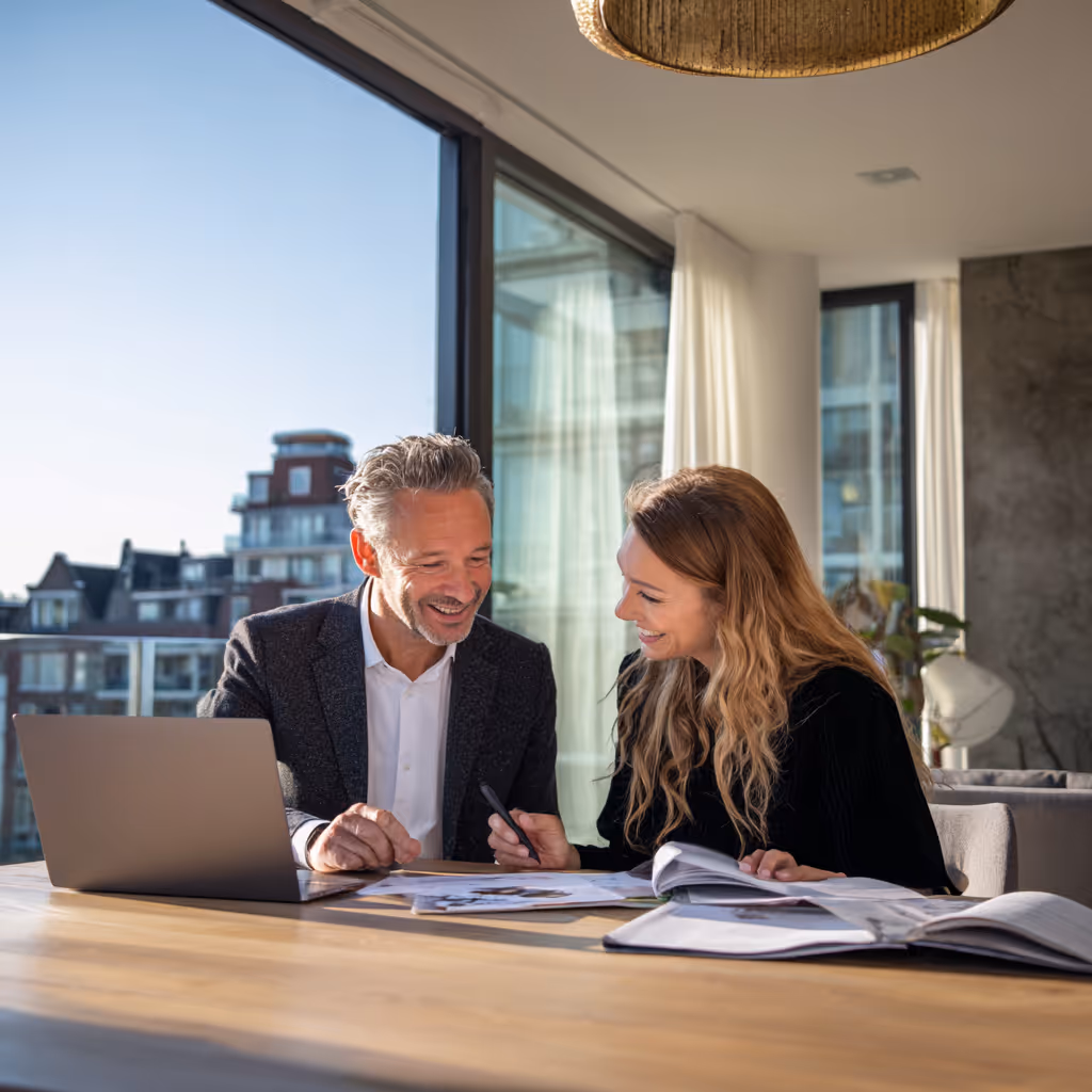Twee mensen die aan een bureau werken, lachend en overleggend met een laptop en open boeken in een lichte ruimte met grote ramen.