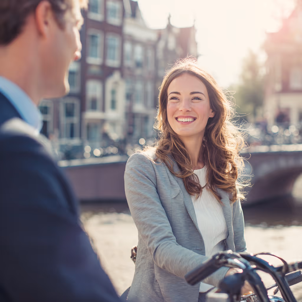 Vrouw lacht terwijl ze naast een man op een fiets staat met grachtenhuisjes op de achtergrond.