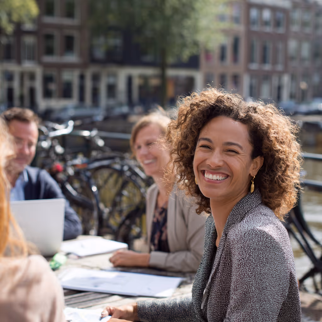 Vrouw met krullend haar lacht zittend aan een tafel buiten met drie andere mensen op een zonnige dag in een stedelijke omgeving.