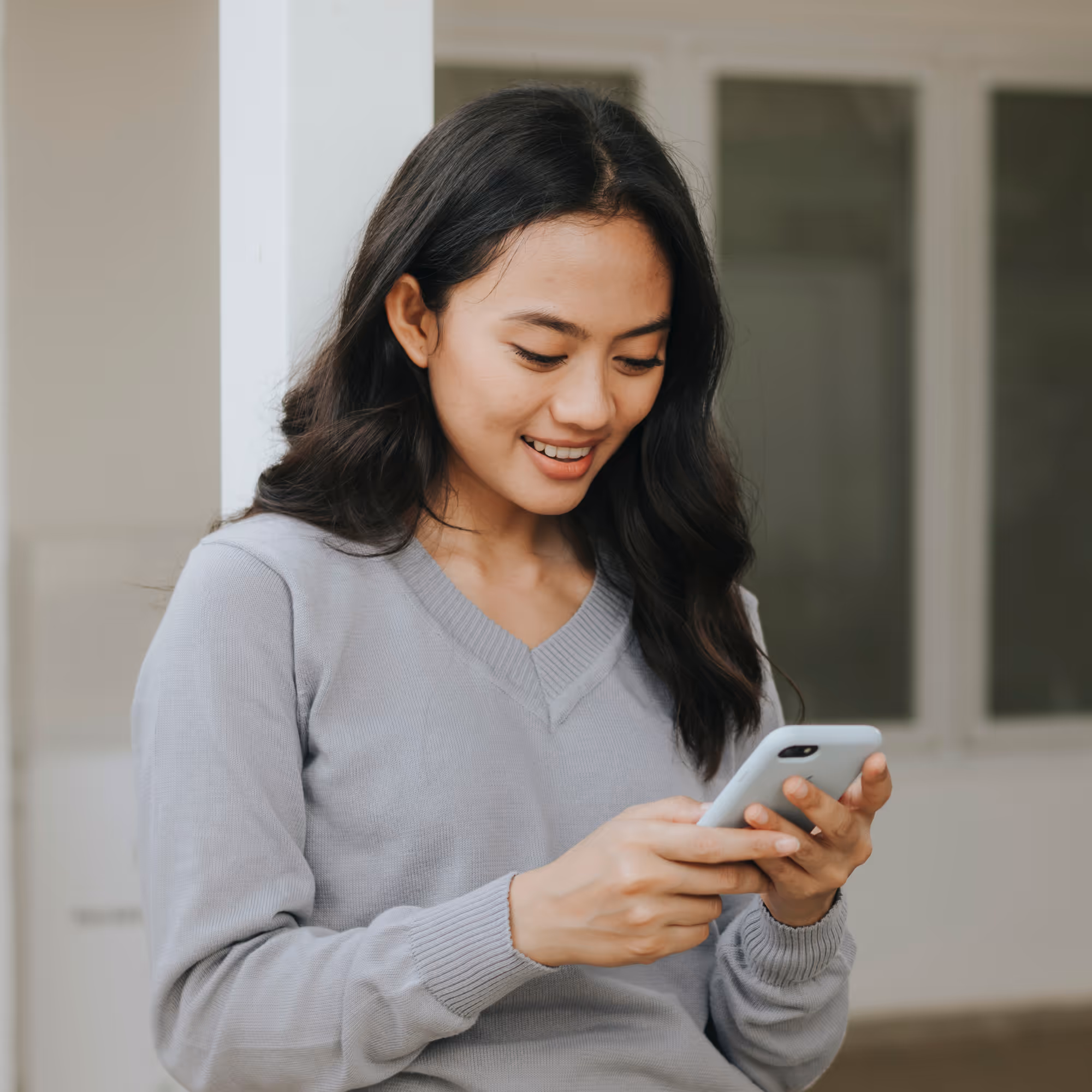 Vrouw met grijze trui glimlachend terwijl ze op haar smartphone kijkt.