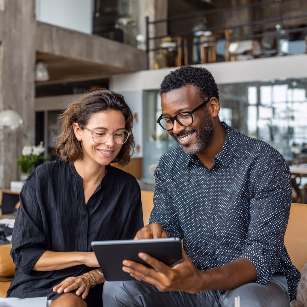 Twee lachende mensen zitten samen en bekijken iets op een tablet in een moderne kantooromgeving.