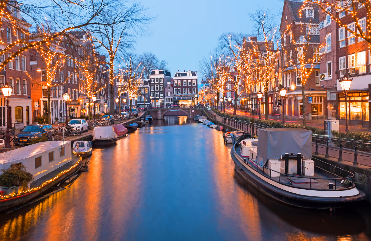 Avondzicht op een Amsterdamse gracht met boten en bomen versierd met lichtjes langs de grachtenpandjes.