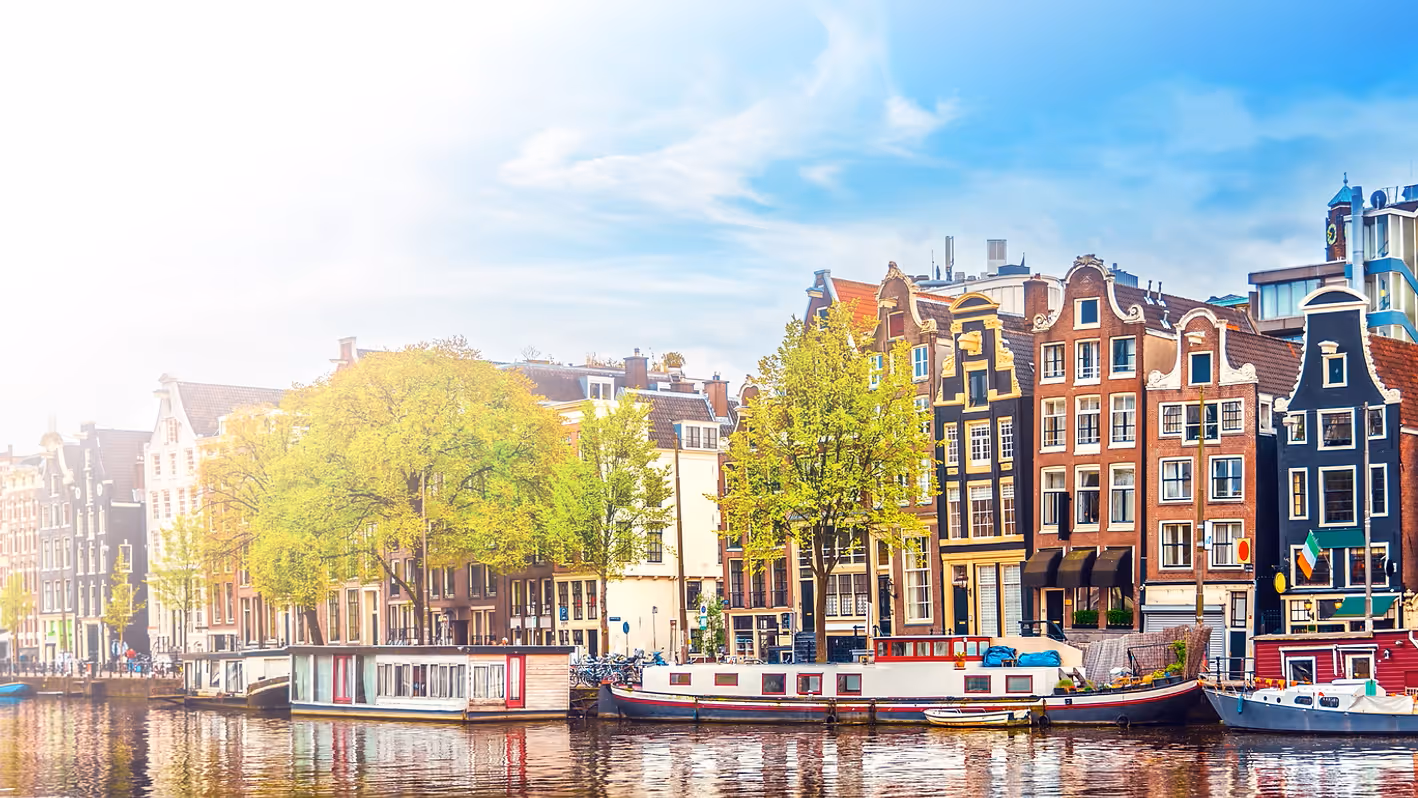 Traditionele smalle huizen langs een gracht met boten en bomen in Amsterdam bij helder weer.