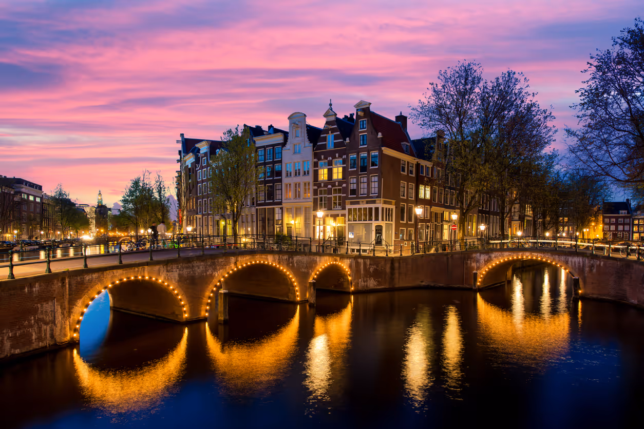 Verlichte bruggen over een gracht met karakteristieke Amsterdamse grachtenpanden bij zonsondergang.