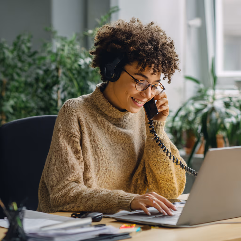 Vrouw met krullend haar en bril die glimlacht terwijl ze met een telefoon spreekt en op een laptop werkt in een kantoorruimte.