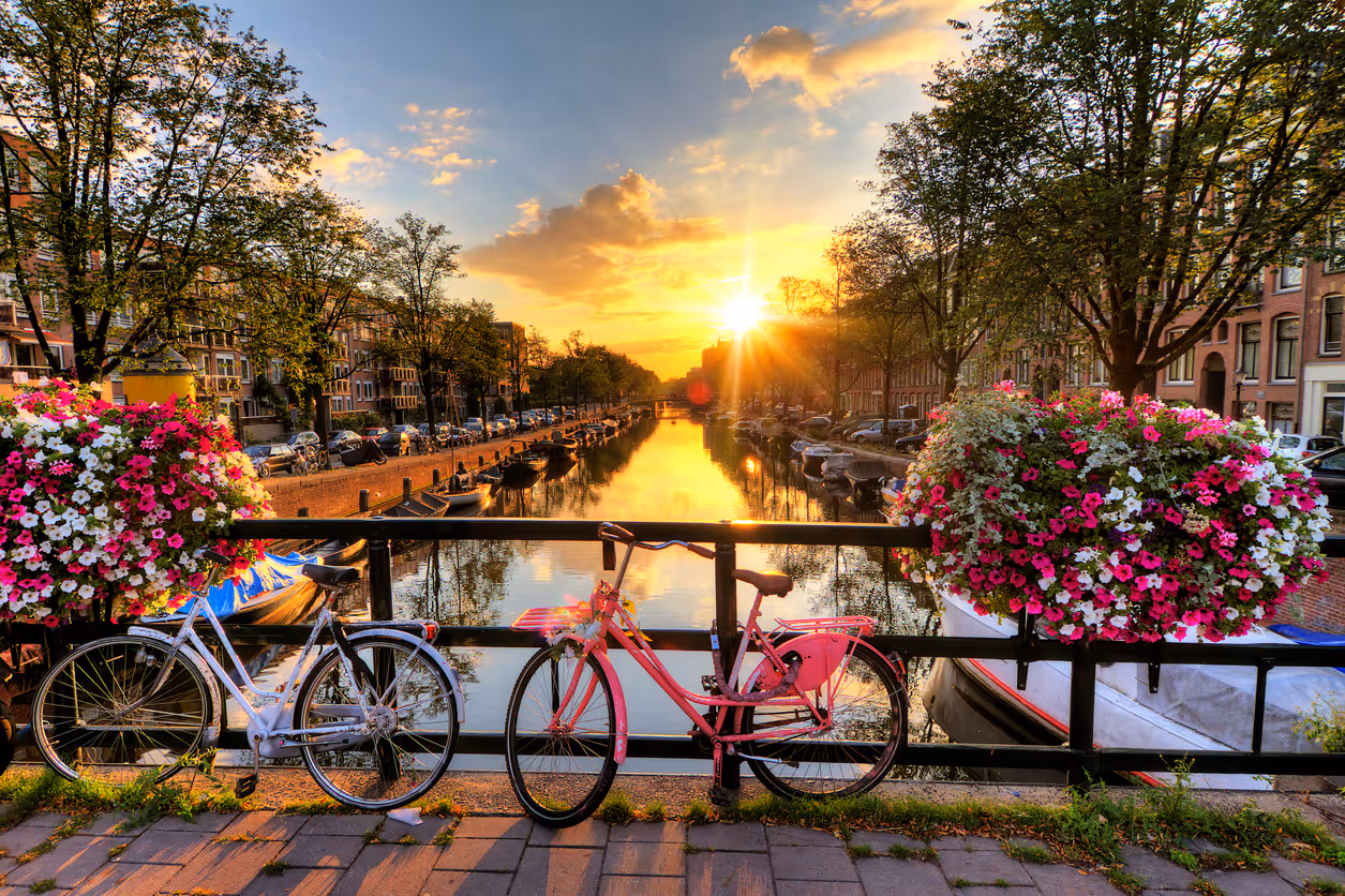 Fietsen vastgemaakt aan een brug met bloemenbakken, uitzicht op een gracht en een zonsondergang in een stad.