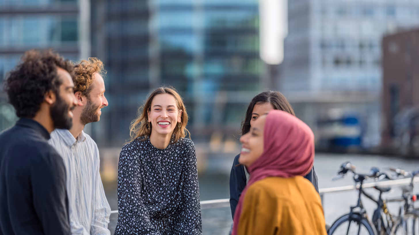 Een groep van vijf diverse jonge volwassenen die buiten lachen en ontspannen bij een waterkant met fietsen op de achtergrond.