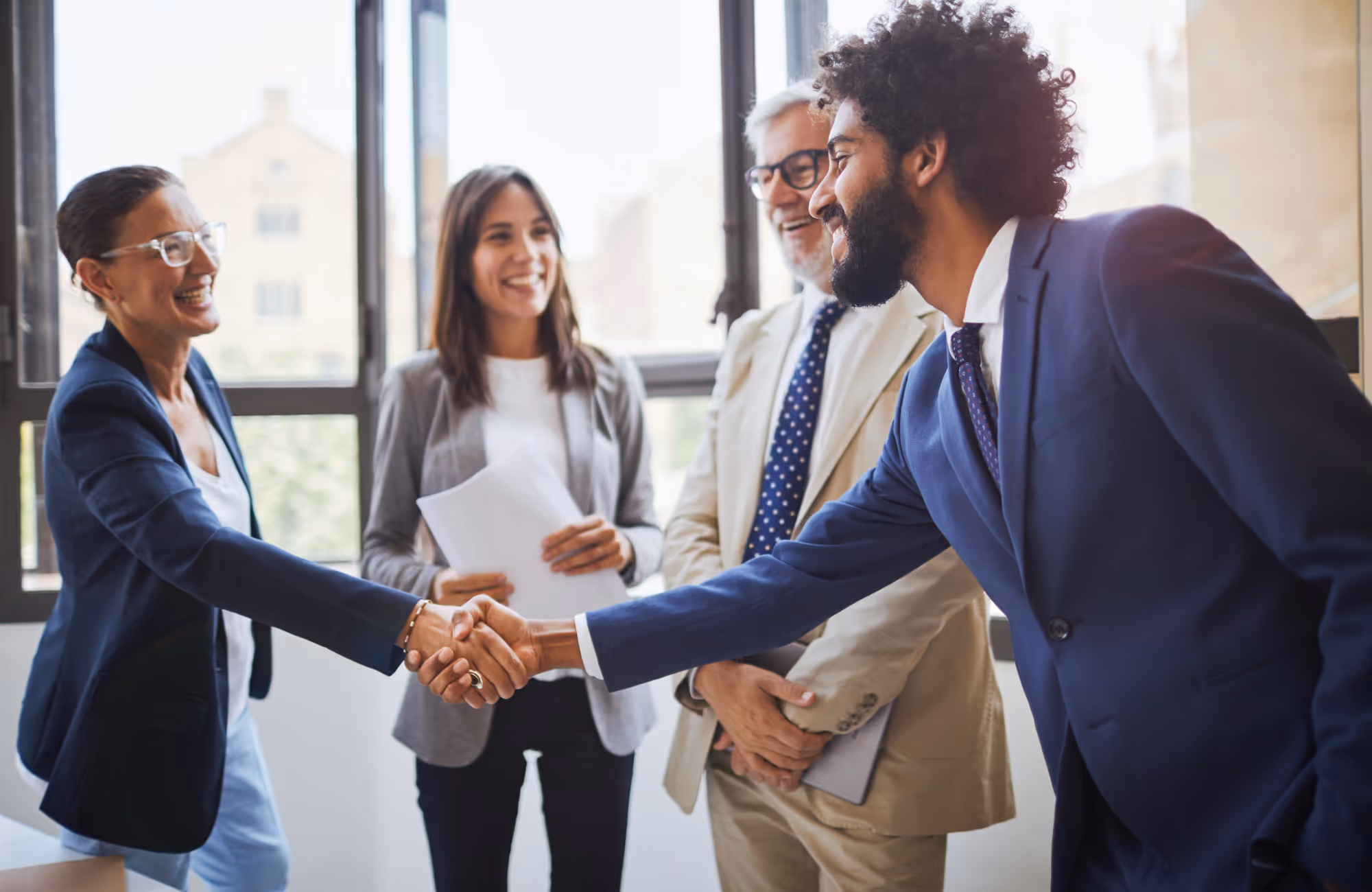 Vier professionals schudden handen en lachen in een goed verlichte kantoorruimte met grote ramen.
