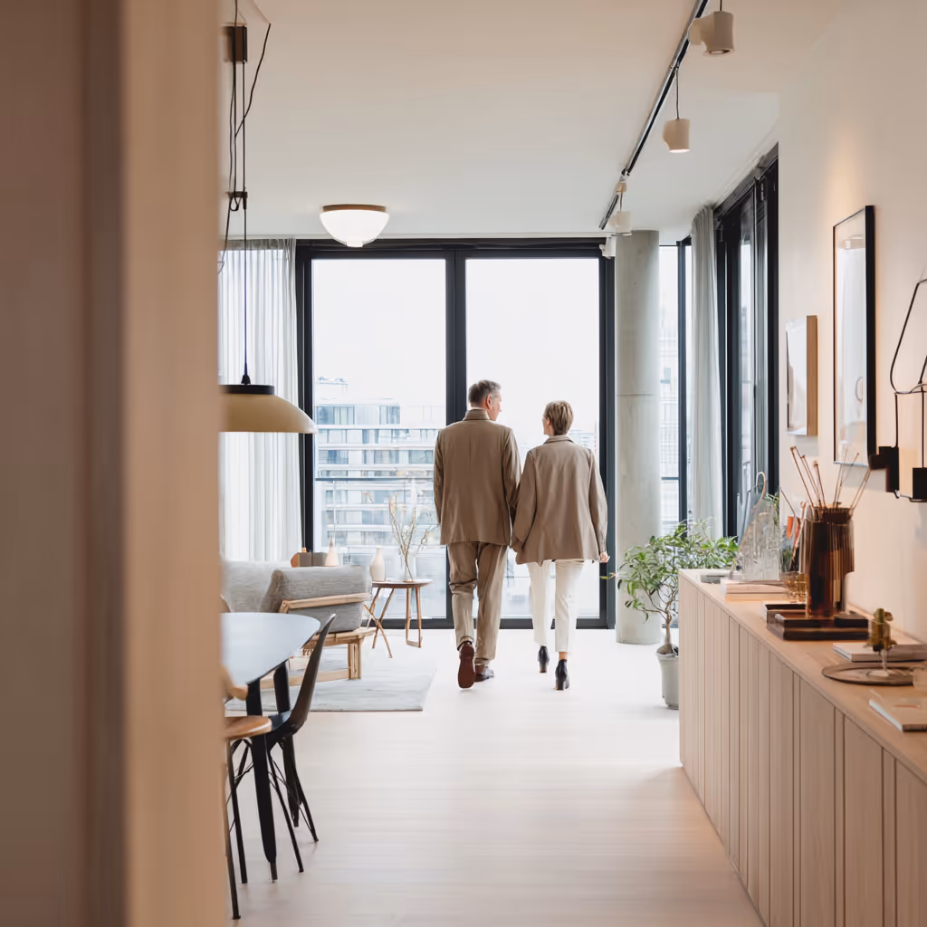 Koppel loopt hand in hand door een modern en licht appartement met grote ramen.