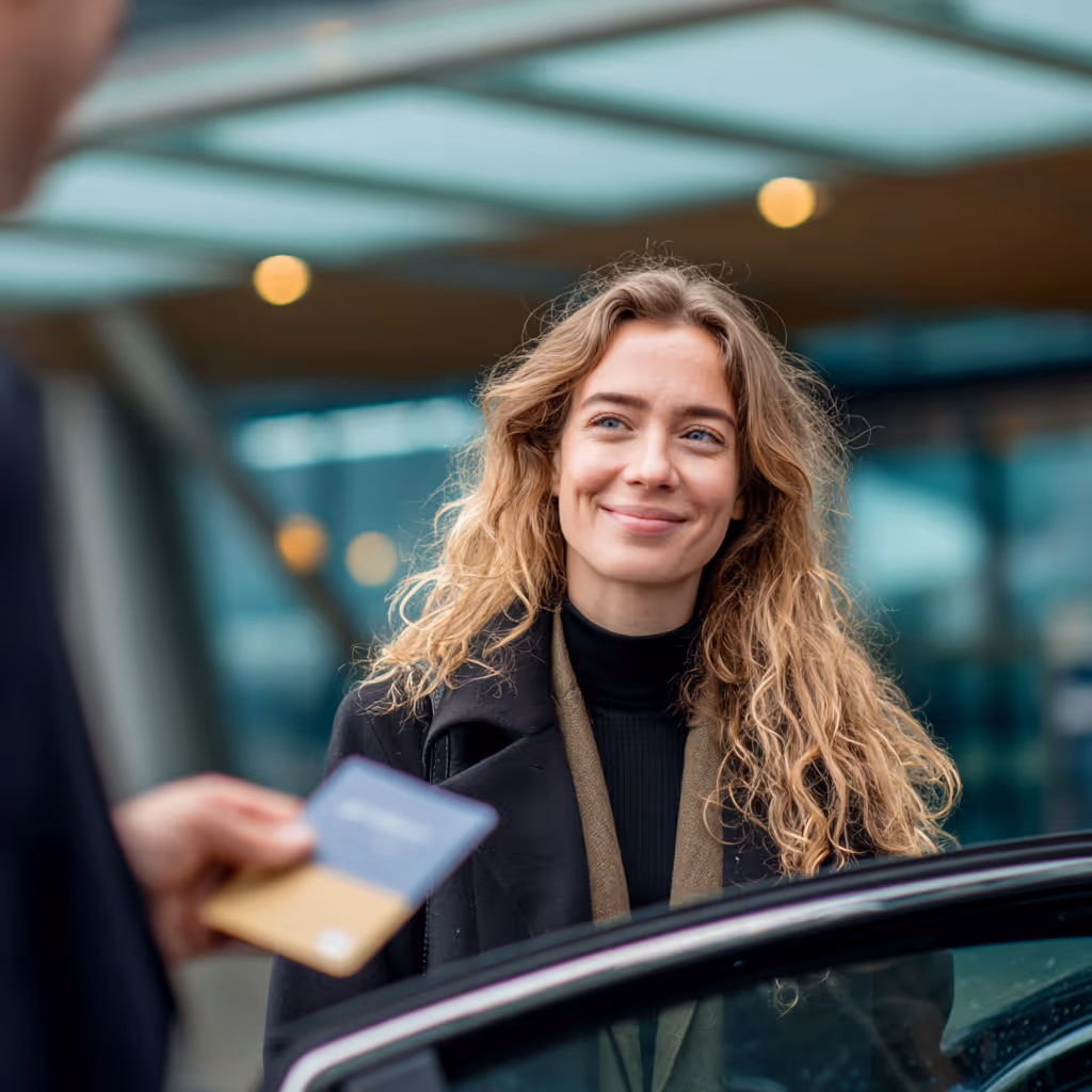 Vrouw met lang blond haar lacht terwijl ze voor de open autodeur staat, een persoon reikt een pas voor haar aan.