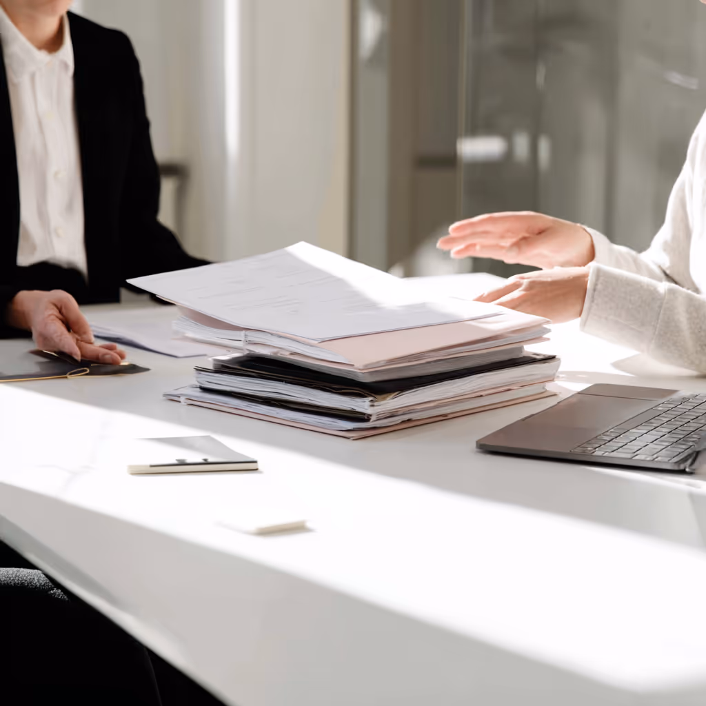 Twee personen zitten aan een tafel met een stapel documenten en een laptop tussen hen in.