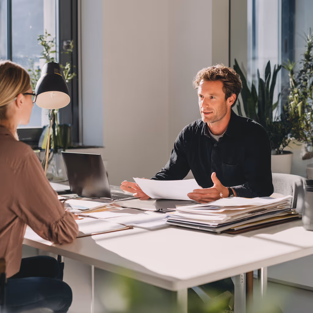 Man praat met vrouw aan tafel in een goed verlichte kantooromgeving met documenten en laptop.