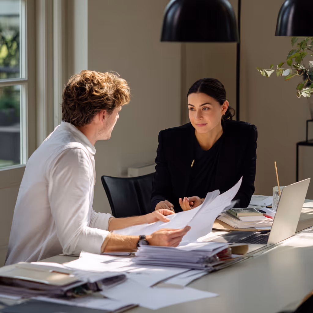 Twee mensen hebben een gesprek aan een bureau met stapels papieren en een open laptop in een kantoorruimte.