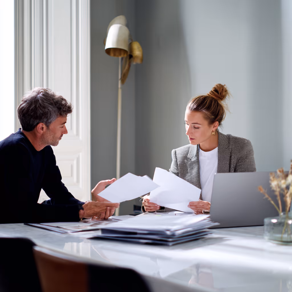 Twee personen zittend aan een tafel die documenten bespreken in een lichte kantoorruimte.