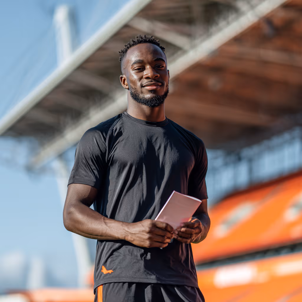 Man in zwart sportshirt en -broek staat buiten in stadion met papieren in zijn handen.