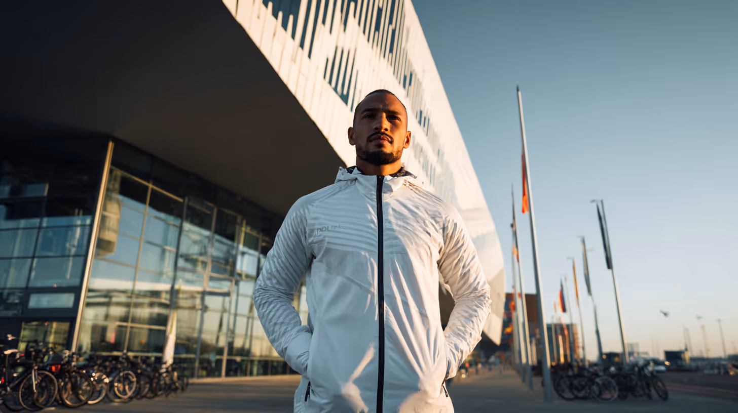 Man in een witte jas staat met handen in zijn zakken voor een modern gebouw met vlaggen en fietsen in de achtergrond bij zonsondergang.